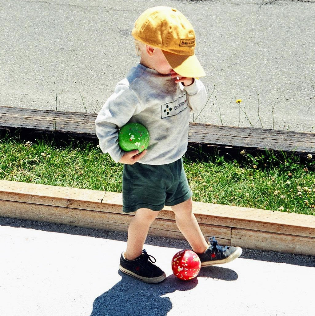Light-skinned toddler in a yellow ball cap standing on a bocce court holding a bocce ball with another ball between his/her feet.